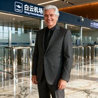 一位五十岁左右的外籍商务男士，银发梳理整齐，穿着深灰色西装，面带微笑站在白云机场贵宾通道前